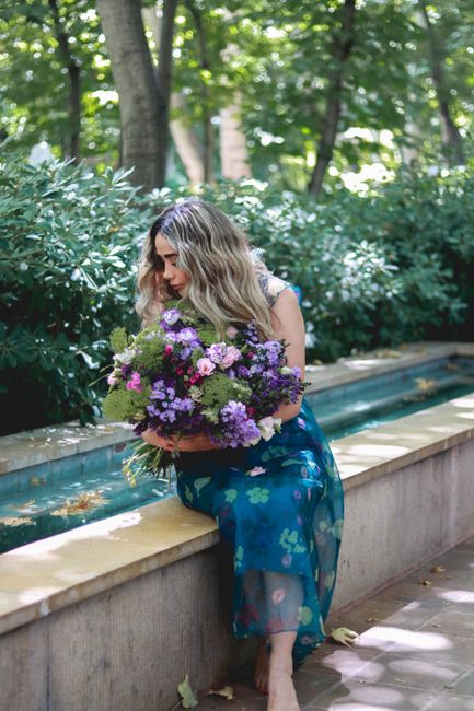 A young woman with wavy, shoulder-length blonde hair is sitting outdoors on a stone ledge beside a low, rectangular water feature, holding a vibrant floral bouquet composed of purple and lavender, gre